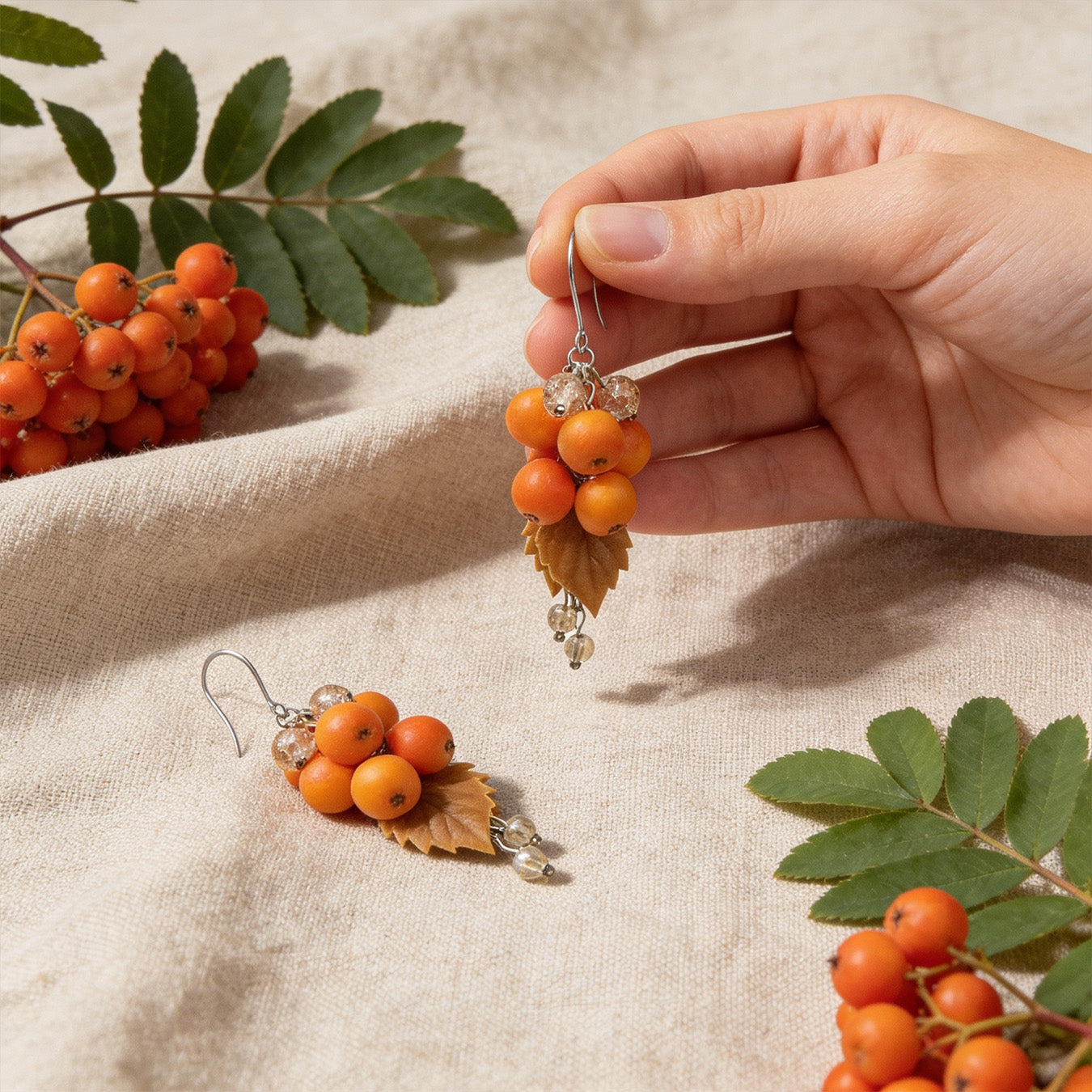 Rowan Berry Earrings photographed with fresh rowan berries and leaves in soft natural light, with one earring gently held by hand for scale. Handmade in Finland by byMinta.
