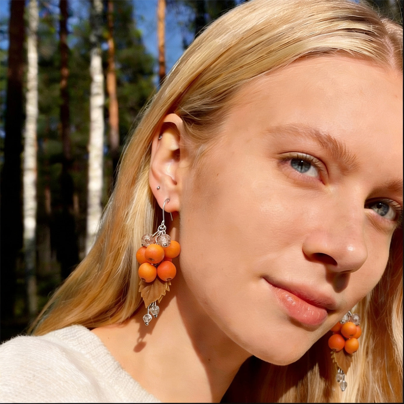 Blonde woman wearing Rowan Berry Earrings in a sunlit Nordic forest. Handmade earrings inspired by the glowing orange-red berries of autumn.