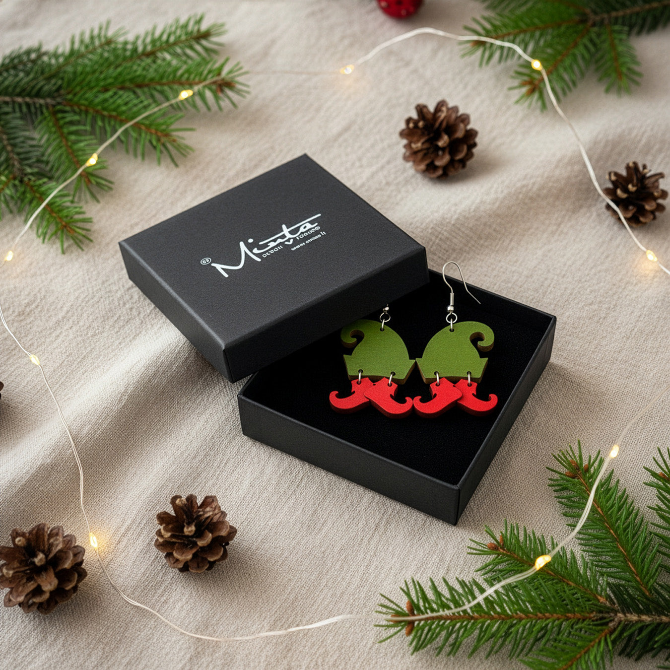 Elf hat earrings presented in a black gift box with spruce branches, pinecones, and soft fairy lights, creating a festive holiday atmosphere. Handmade in Finland by byMinta.