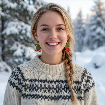 Smiling blonde woman wearing green and red elf hat earrings in a snowy winter landscape, dressed in a cozy Nordic knit sweater. Handmade in Finland by byMinta.