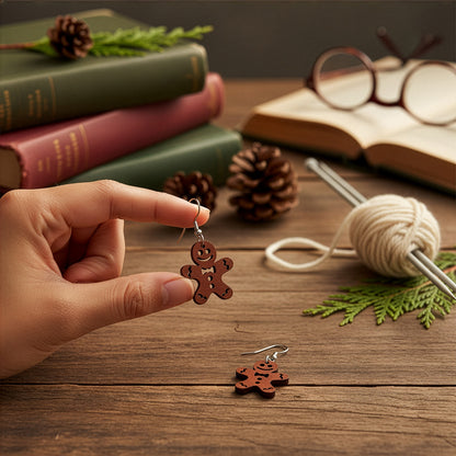 Hand holding a gingerbread cookie earring on a wooden table with books, knitting needles, yarn, and pinecones in a cozy winter setting. Handmade in Finland by byMinta.