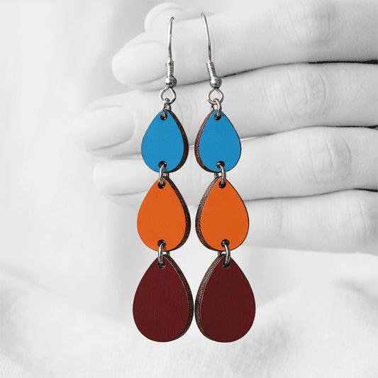 Walnut, orange and skyblue Sadetta (Rain) plywood earrings made in Finland, featuring intricate laser-cut patterns and silver-toned hooks, photographed hanging from a hand against a softly blurred light linen background.