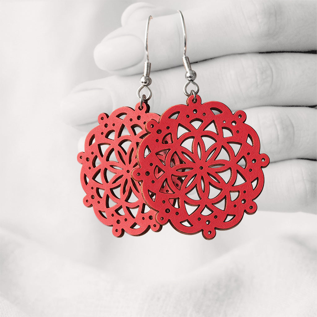 Red Pitsimandala plywood earrings made in Finland, featuring intricate laser-cut patterns and silver-toned hooks, photographed hanging from a hand against a softly blurred light linen background.