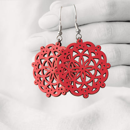 Red Pitsimandala plywood earrings made in Finland, featuring intricate laser-cut patterns and silver-toned hooks, photographed hanging from a hand against a softly blurred light linen background.
