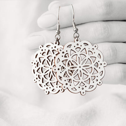 White Pitsimandala plywood earrings made in Finland, featuring intricate laser-cut patterns and silver-toned hooks, photographed hanging from a hand against a softly blurred light linen background.
