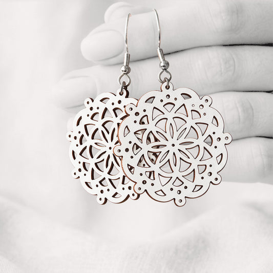 White Pitsimandala plywood earrings made in Finland, featuring intricate laser-cut patterns and silver-toned hooks, photographed hanging from a hand against a softly blurred light linen background.