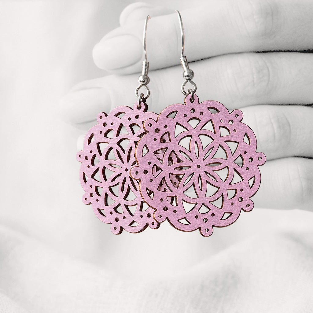 Mauve Pitsimandala plywood earrings made in Finland, featuring intricate laser-cut patterns and silver-toned hooks, photographed hanging from a hand against a softly blurred light linen background.
