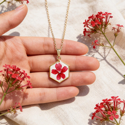 Hand holding a handmade botanical necklace for size reference, featuring a real pressed red verbena flower on a white background. Surrounded by vibrant red verbena flowers and soft linen in bright natural daylight with a warm summer atmosphere.