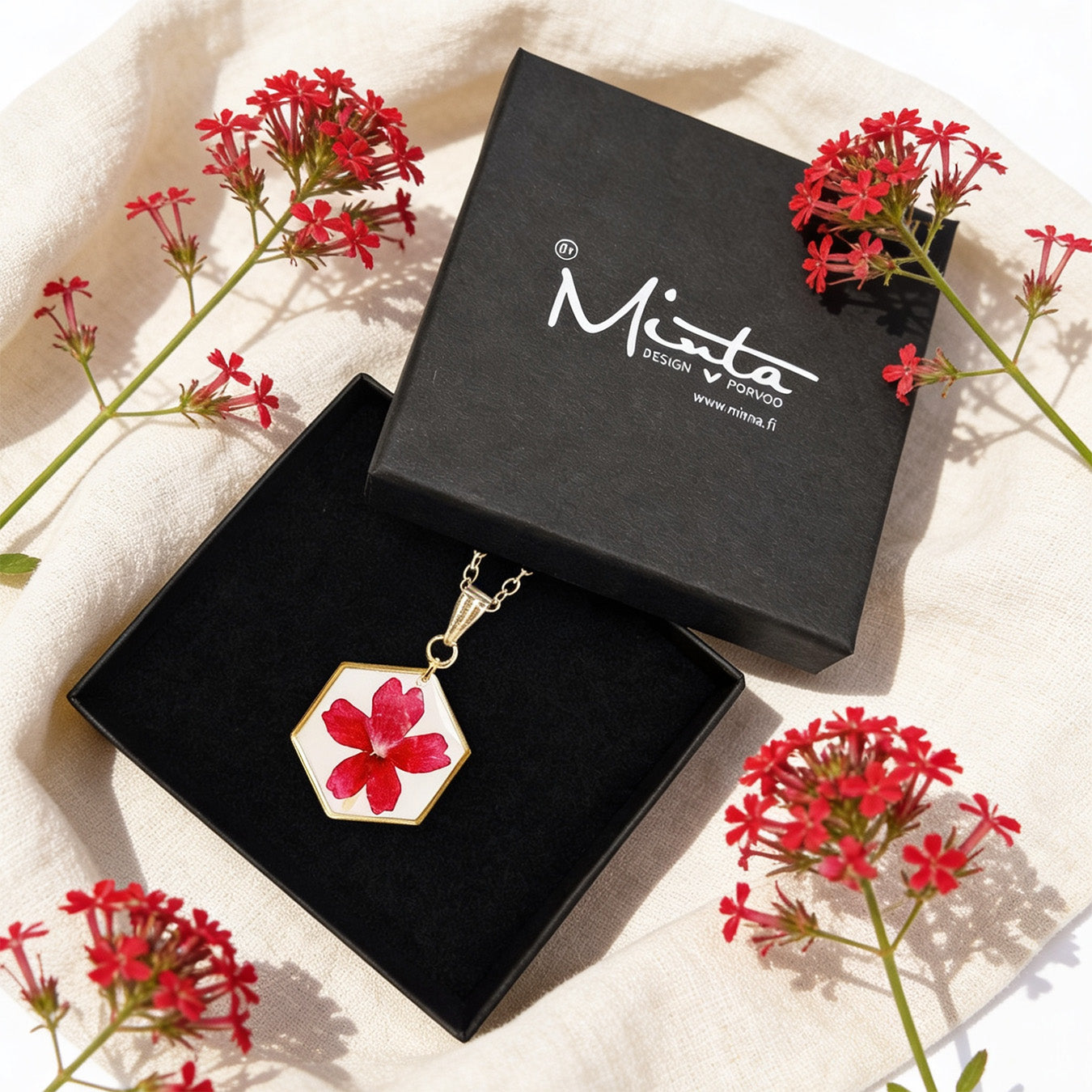 Handmade botanical necklace with a real pressed red verbena flower displayed in a black byMinta jewelry box. Surrounded by vibrant red verbena blooms and soft linen in bright natural daylight with a warm and elegant summer atmosphere.