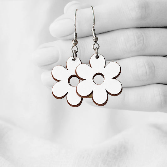 White Retro Flower plywood earrings made in Finland, photographed hanging from a hand in black and white against a softly blurred light background.