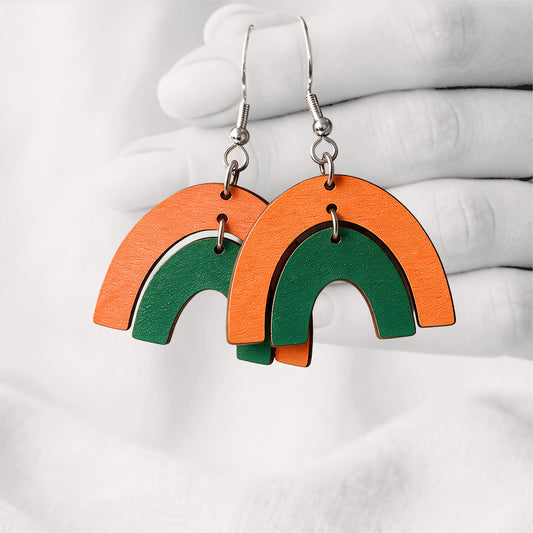 Orange and green Riemukaari Duo plywood earrings handmade in Finland, shown against a softly blurred hand and light linen background
