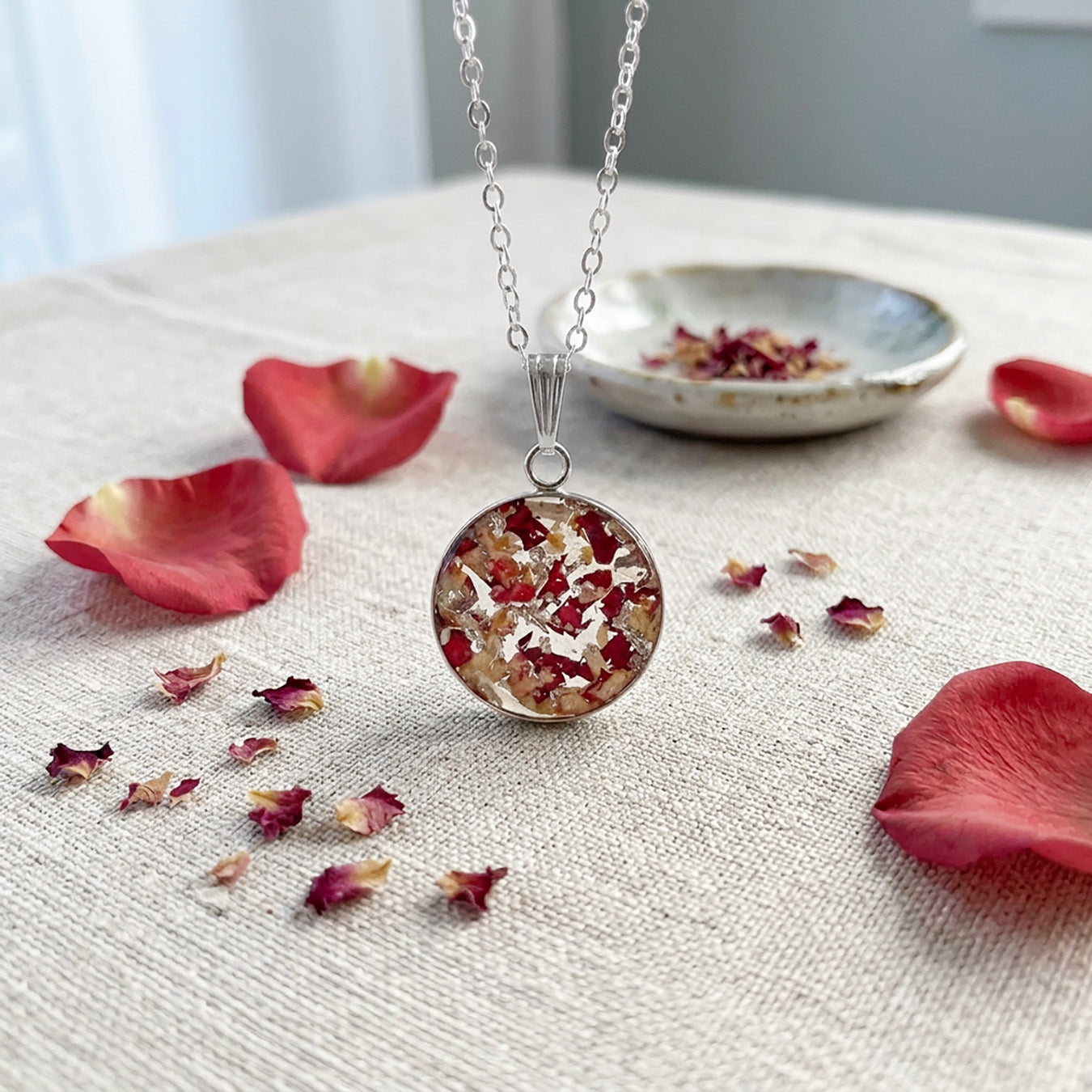 Handmade resin necklace with real rose petal fragments and silver leaf flakes displayed on natural linen fabric surrounded by scattered rose petals.