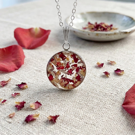 Handmade resin necklace with real rose petal fragments and silver leaf flakes displayed on natural linen fabric surrounded by scattered rose petals.
