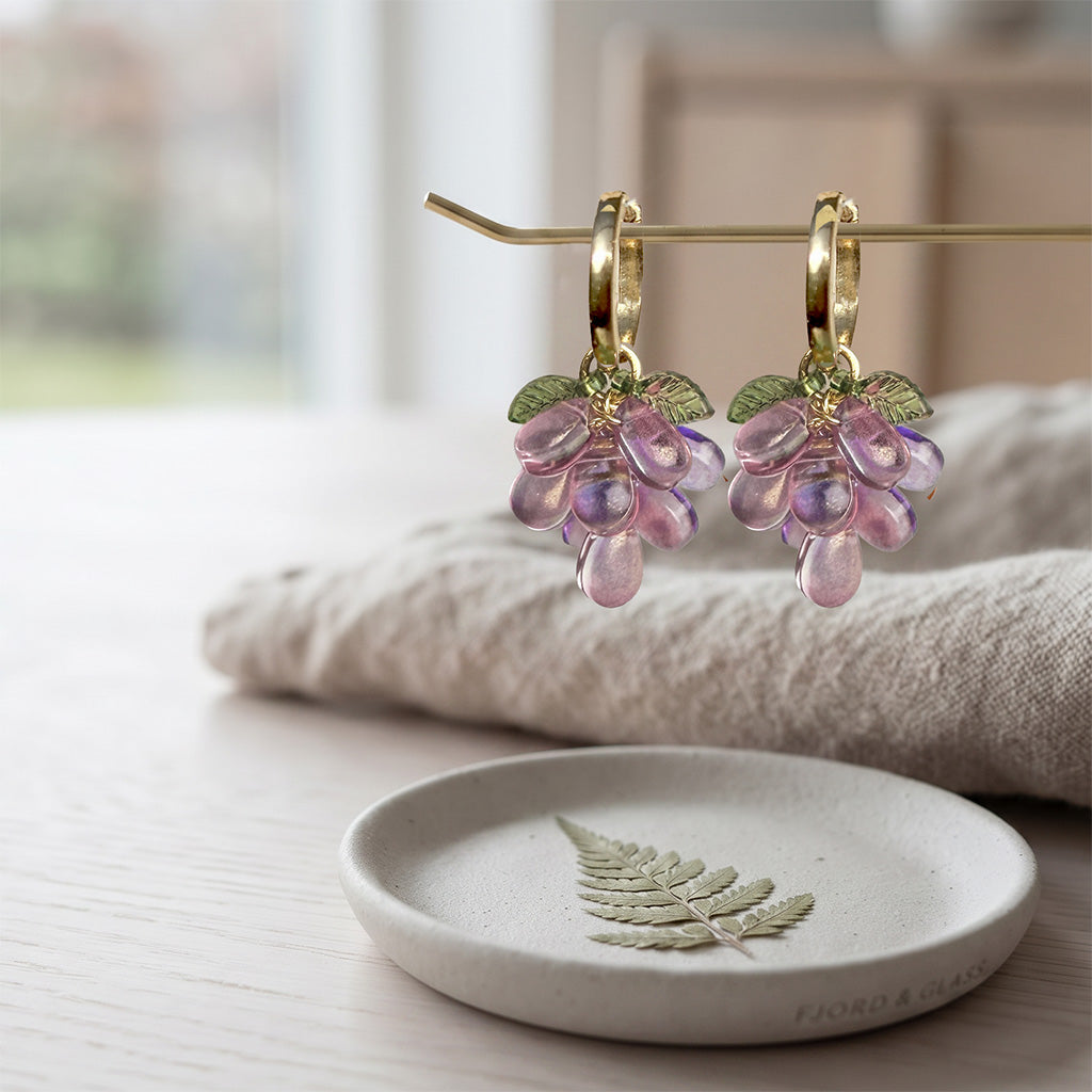 Plum Cluster Earrings with translucent purple glass beads, detachable charm and gold-plated huggie hoops on a soft linen background. Handcrafted in Finland.