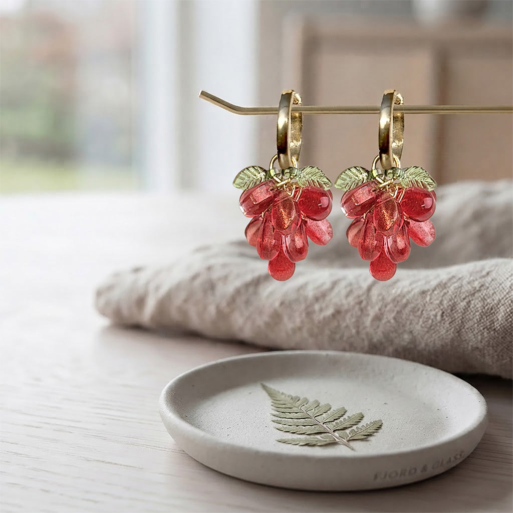 Red Currant Cluster Earrings with translucent red glass beads, detachable charm and gold-plated huggie hoops on a soft linen background. Handcrafted in Finland.