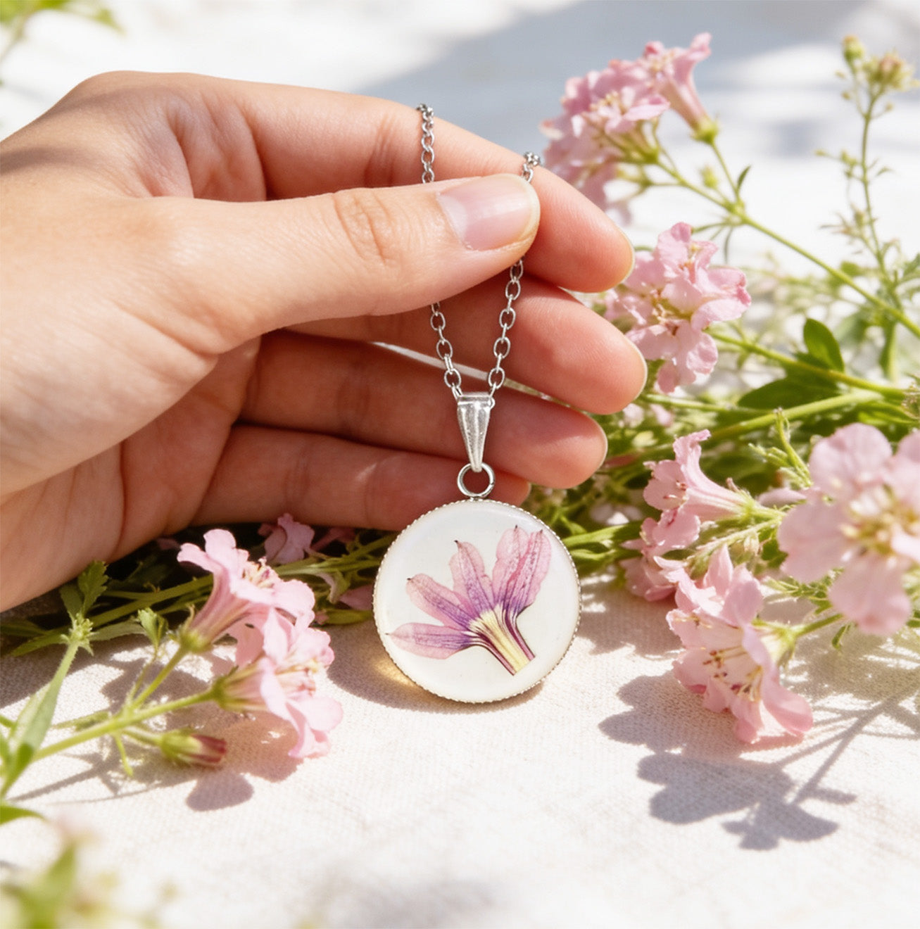 Hand holding a handmade resin necklace with a real pink fan flower (Scaevola) for size reference, surrounded by delicate pink fan flowers in bright natural daylight. Botanical jewelry photographed with a soft and airy summer atmosphere.