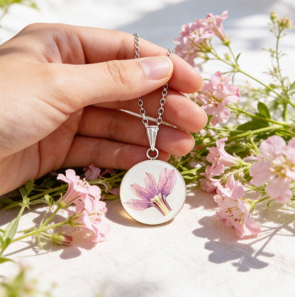 Hand holding a handmade resin necklace with a real pink fan flower (Scaevola) for size reference, surrounded by delicate pink fan flowers in bright natural daylight. Botanical jewelry photographed with a soft and airy summer atmosphere.