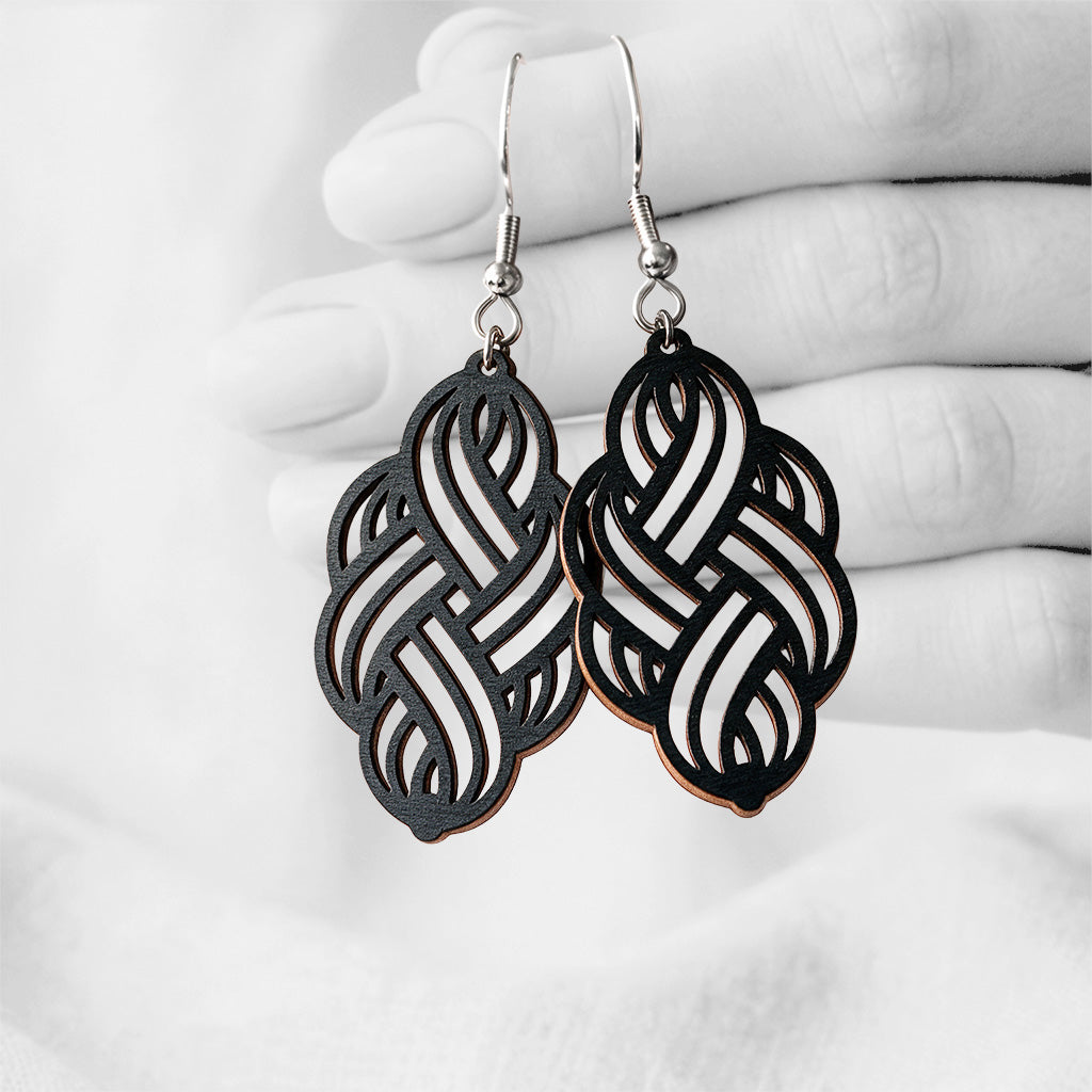Black Solmu (Knot) plywood earrings handmade in Finland, photographed hanging against a black-and-white hand background.