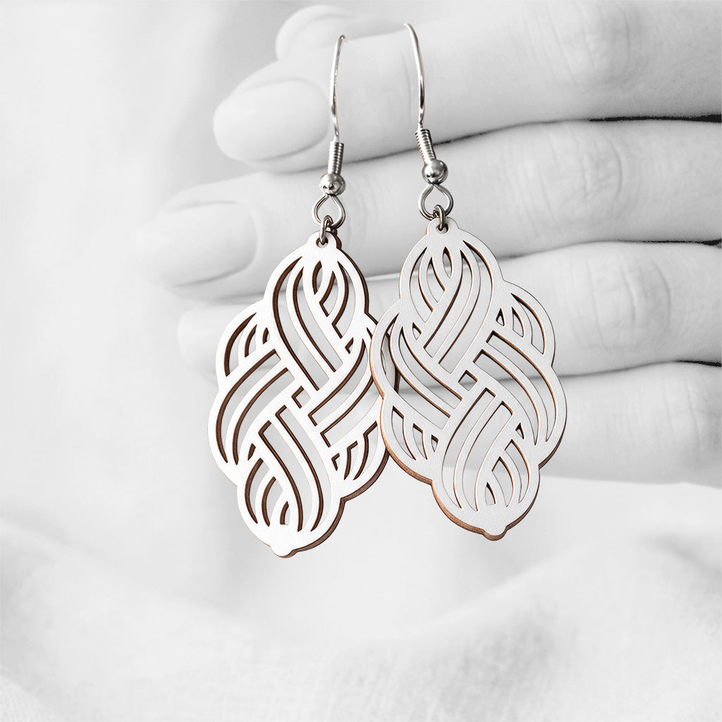 White Solmu (Knot) plywood earrings handmade in Finland, photographed hanging against a black-and-white hand background.
