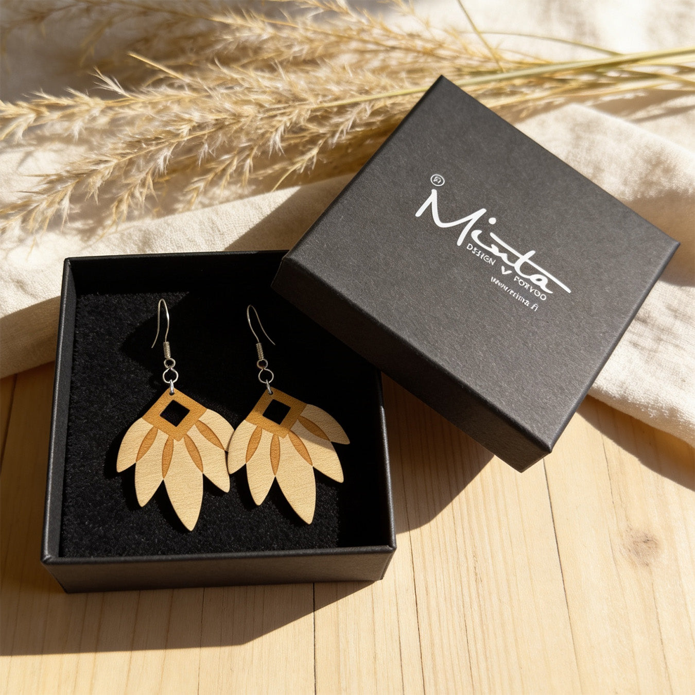 Handmade wooden leaf earrings displayed in a black jewelry box on a light wooden surface with soft linen fabric and dried grasses. Warm natural sunlight creates a calm Nordic atmosphere with elegant natural styling.
