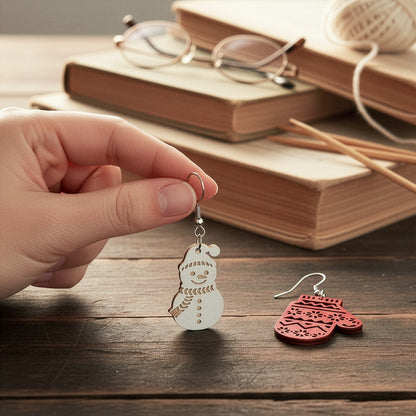 Hand holding the snowman earring on a wooden table with books, glasses, knitting needles, and yarn, with the mitten earring beside it. Handmade in Finland by byMinta.