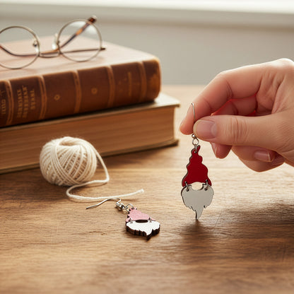 Hand holding a Christmas gnome earring on a wooden table with vintage books, glasses, and soft white yarn in a warm winter setting. Handmade in Finland by byMinta.
