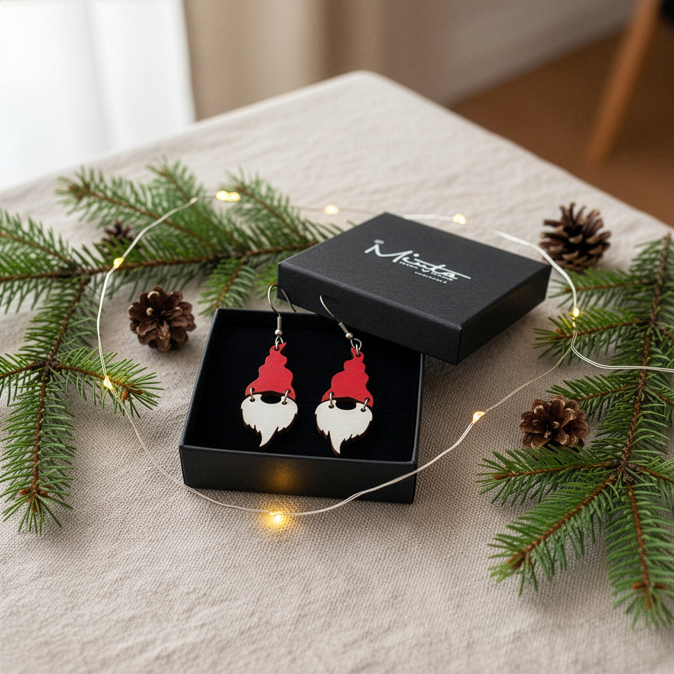 Christmas gnome earrings presented in a gift box with spruce branches, pinecones, and warm fairy lights, creating a cozy holiday atmosphere. Handmade in Finland by byMinta.