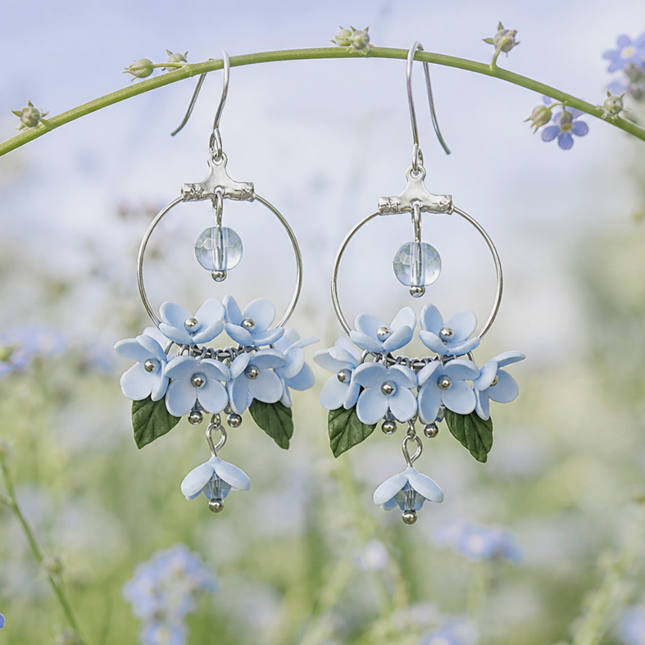 Mist Flower Hoop Earrings displayed hanging from a flower stem in a dreamy summer meadow. Handmade in Finland by byMinta.