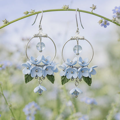 Mist Flower Hoop Earrings displayed hanging from a flower stem in a dreamy summer meadow. Handmade in Finland by byMinta.