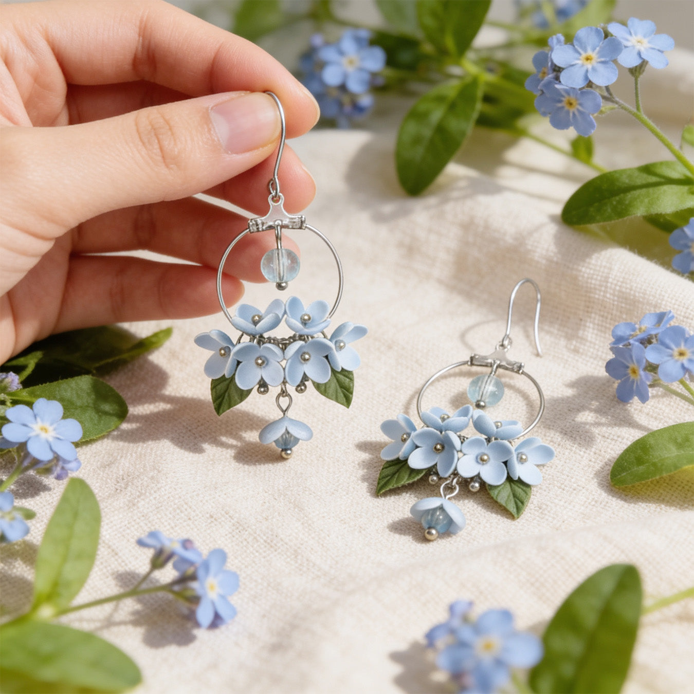 Mist Flower Hoop Earrings photographed with forget-me-nots in soft natural light, with one earring gently held by hand for scale. Handmade in Finland by byMinta.