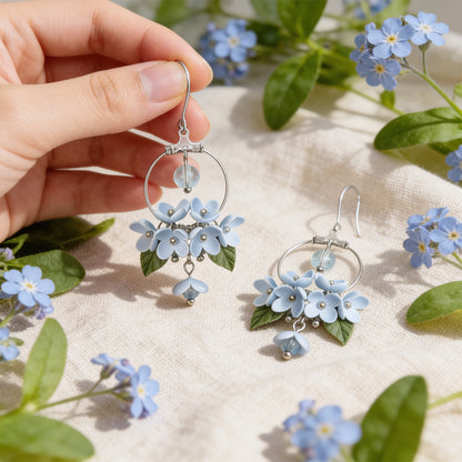 Mist Flower Hoop Earrings photographed with forget-me-nots in soft natural light, with one earring gently held by hand for scale. Handmade in Finland by byMinta.