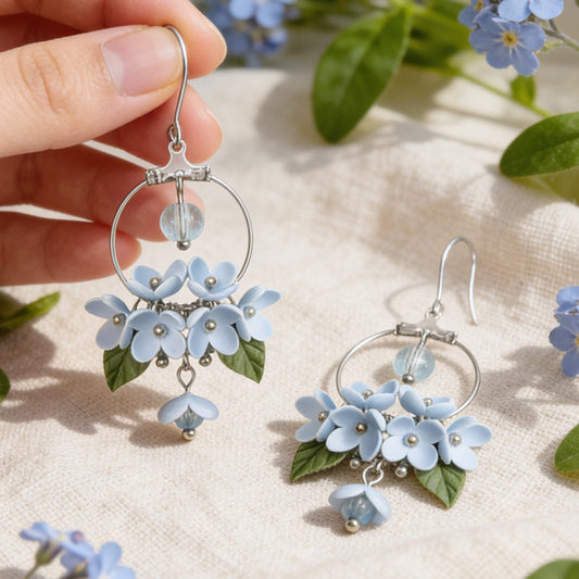 Mist Flower Hoop Earrings photographed with forget-me-nots in soft natural light, with one earring gently held by hand for scale. Handmade in Finland by byMinta.