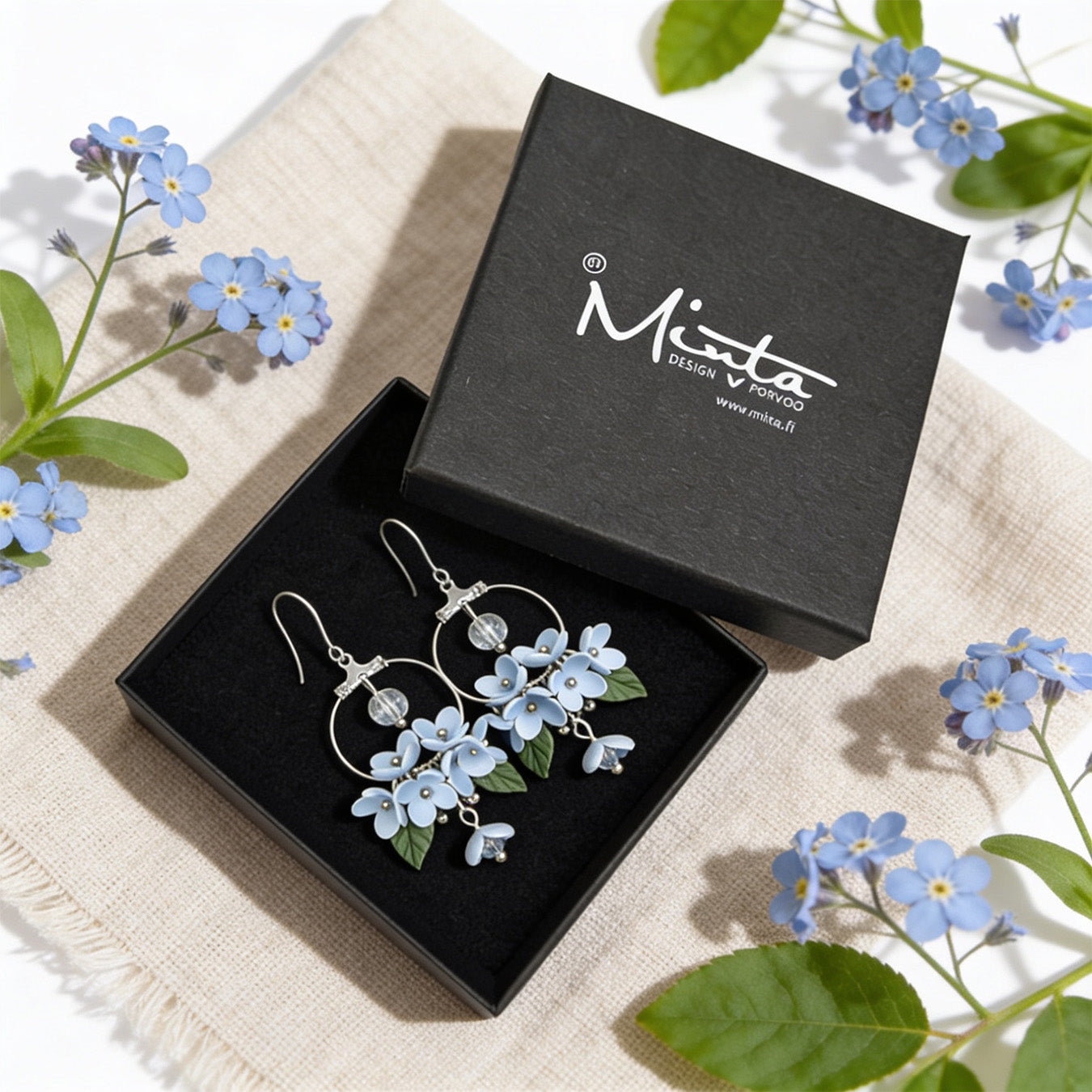 Mist Flower Hoop Earrings presented in a gift box, surrounded by natural linen fabric and delicate blue flowers. Handmade in Finland by byMinta.