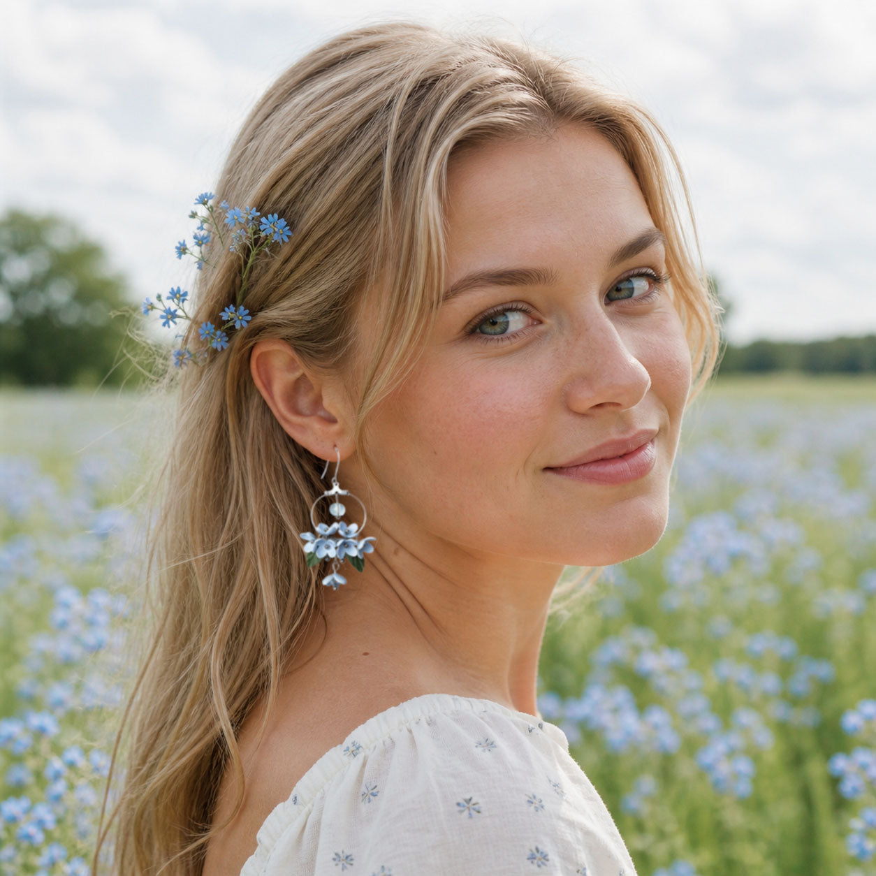 Blonde woman wearing Mist Flower Hoop Earrings in a blooming field of forget-me-nots. Handmade floral earrings inspired by the soft blue tones of a Finnish summer sky.