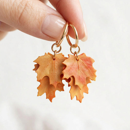 Orange Maple Leaf earrings handmade in Finland, held between fingers against a soft, light background in natural daylight.