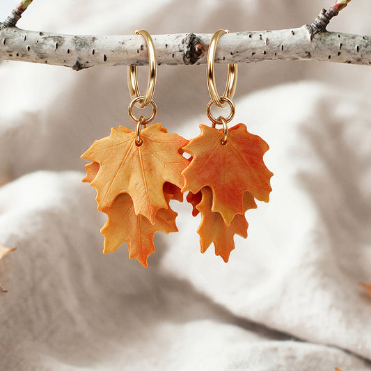 Orange Vaahteranlehti (Maple Leaf) earrings with gold-tone huggie hoops, handcrafted in Finland, hanging from a birch branch against a natural linen background.