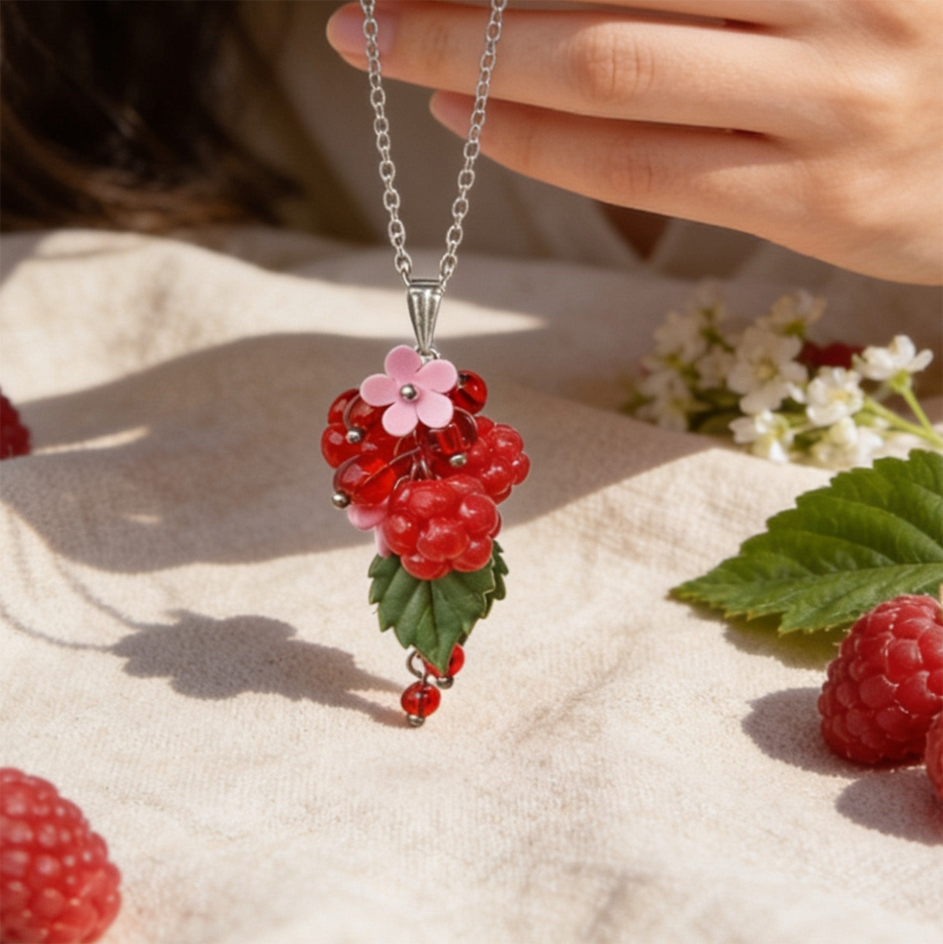 Raspberry Garden Necklace held gently by hand, featuring hand-shaped raspberries and leaves made from polymer clay. Surrounded by natural linen fabric, fresh raspberries, green leaves, and delicate white flowers in soft natural light. Handmade in Finland by byMinta.