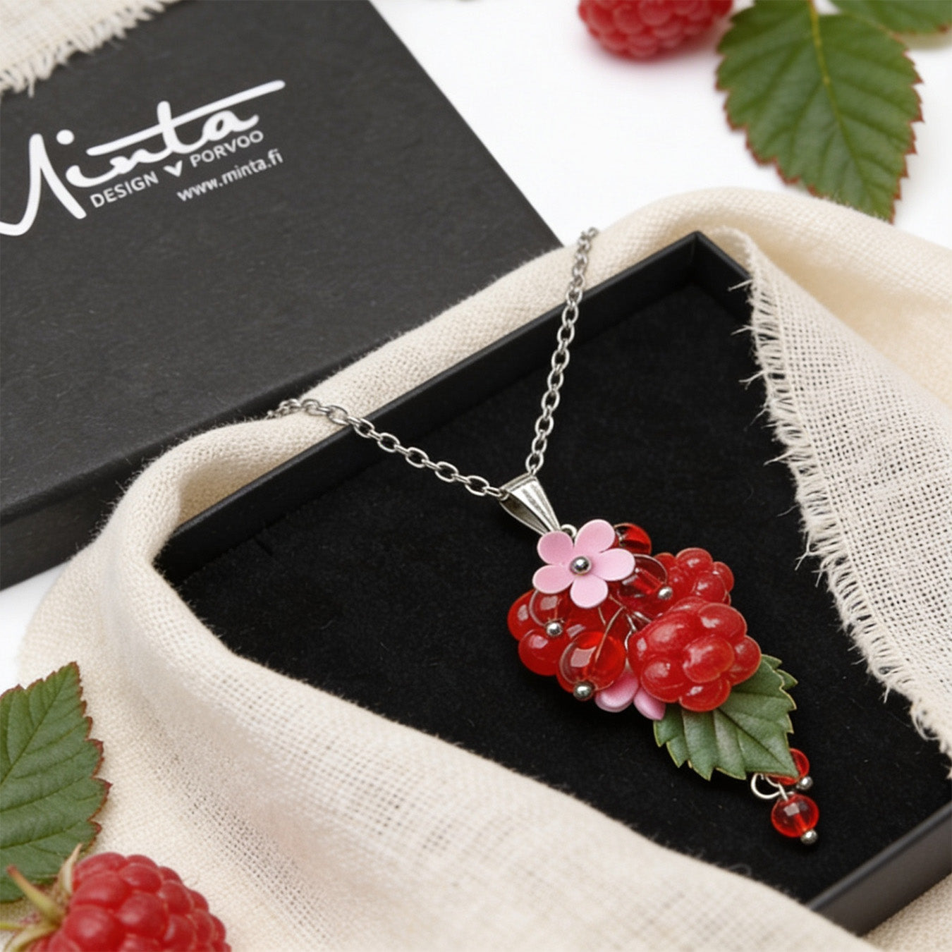Raspberry Garden Necklace presented in a gift box, featuring hand-shaped berries and leaves, surrounded by natural linen fabric, fresh raspberries, green leaves, and delicate white flowers in soft natural light. Handmade in Finland by byMinta.