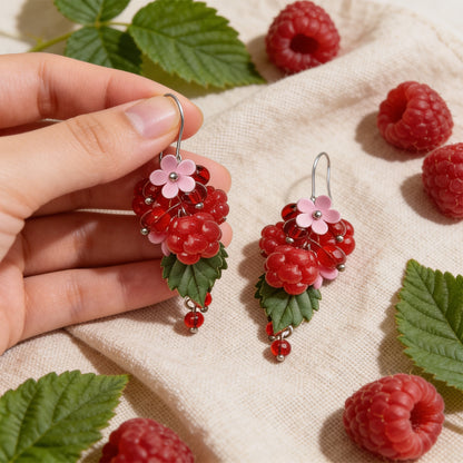 Raspberry Garden Earrings photographed with fresh raspberries and green leaves in soft natural light, with one earring gently held by hand for scale. Handmade in Finland by byMinta.
