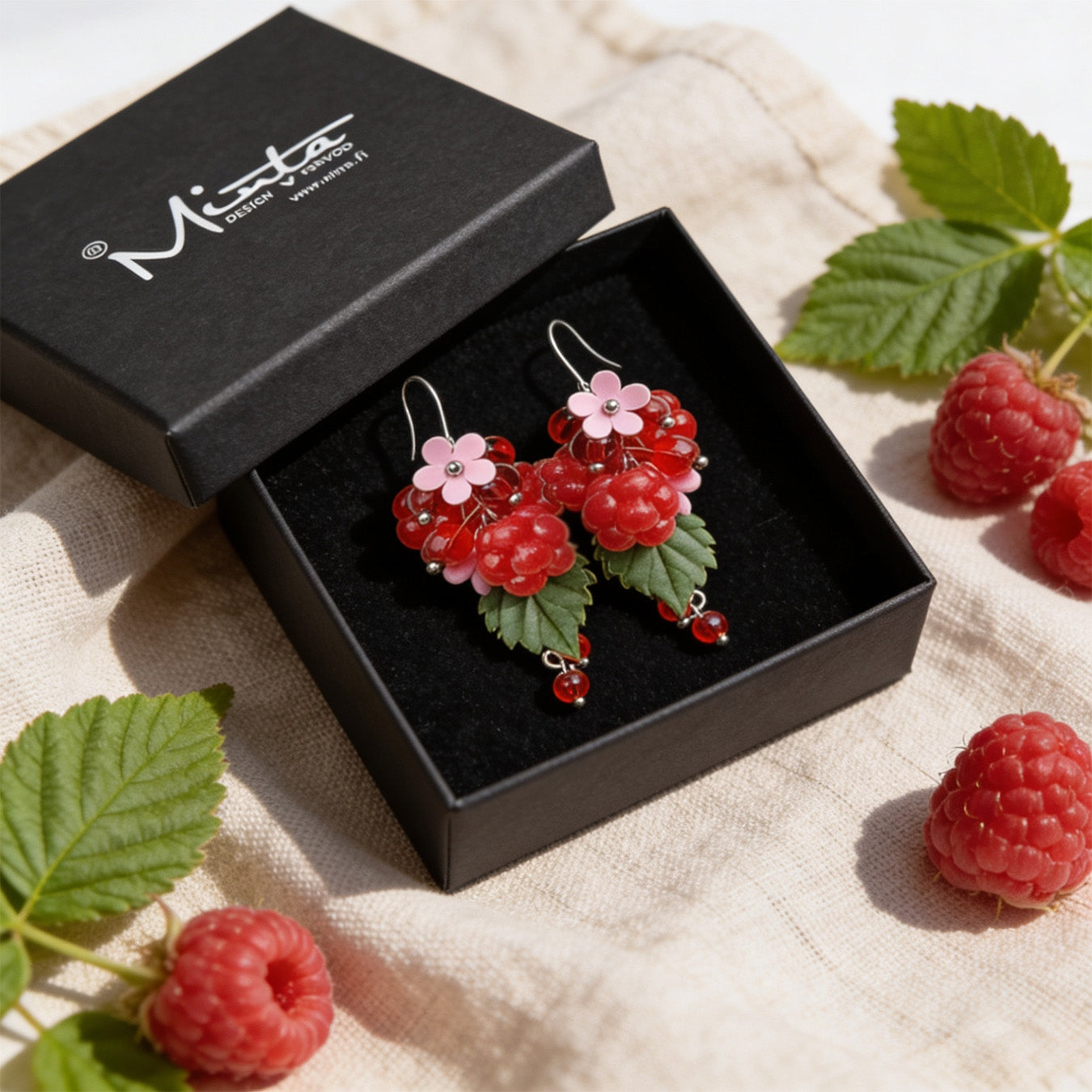 Raspberry Garden Earrings presented in a gift box, surrounded by natural linen fabric, fresh raspberries, and raspberry leaves. Handmade in Finland by byMinta.