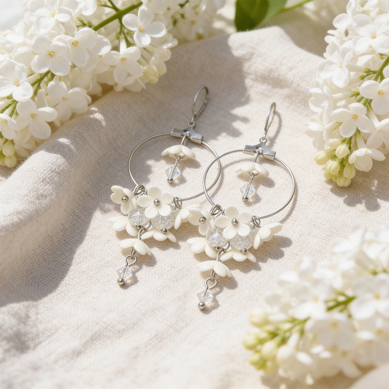 White Lilac Hoop Earrings resting on linen among blooming white lilacs in bright natural daylight. Handmade in Finland by byMinta.