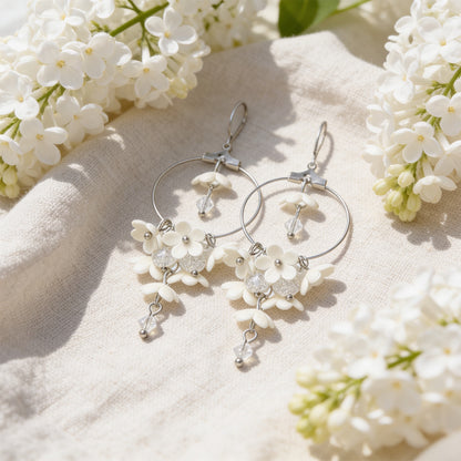 White Lilac Hoop Earrings resting on linen among blooming white lilacs in bright natural daylight. Handmade in Finland by byMinta.