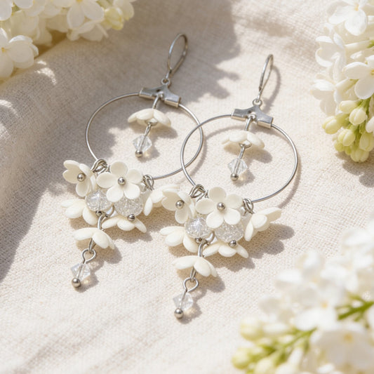 White Lilac Hoop Earrings resting on linen among blooming white lilacs in bright natural daylight. Handmade in Finland by byMinta.