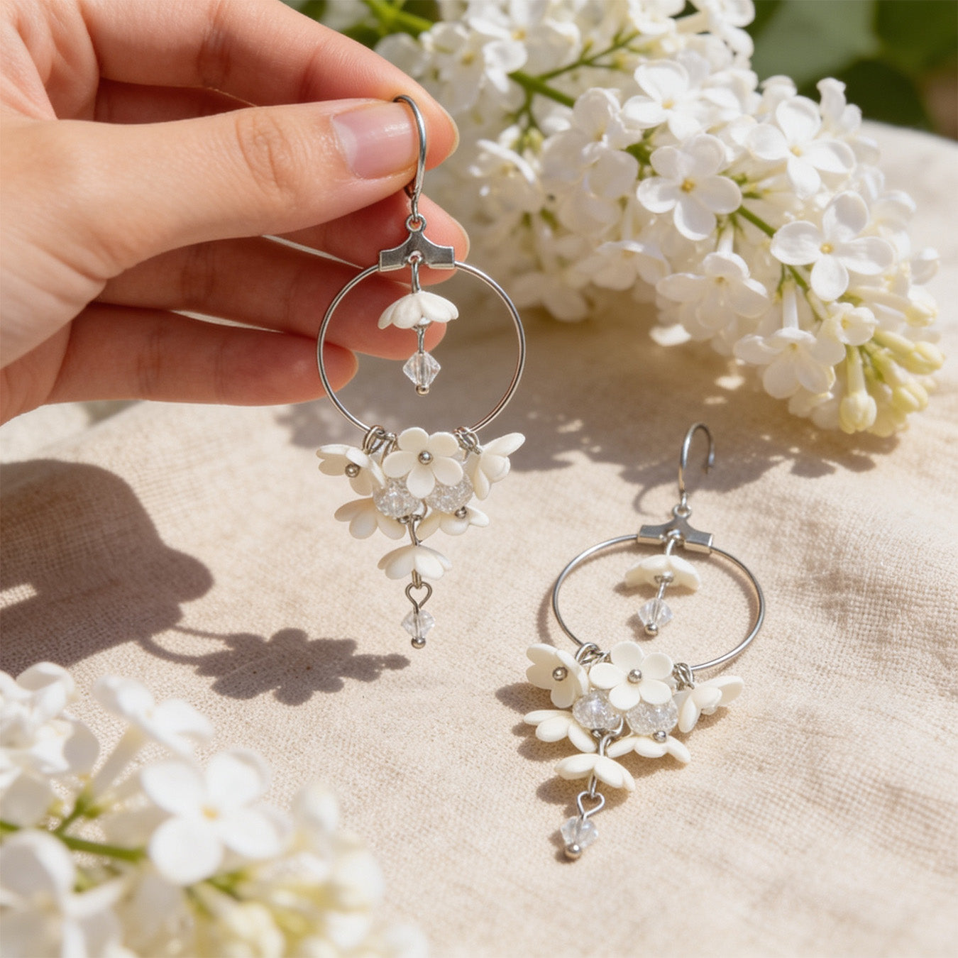 White Lilac Hoop Earrings photographed with blooming white lilacs in soft natural light, with one earring gently held by hand for scale. Handmade in Finland by byMinta.