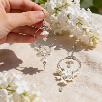 White Lilac Hoop Earrings photographed with blooming white lilacs in soft natural light, with one earring gently held by hand for scale. Handmade in Finland by byMinta.