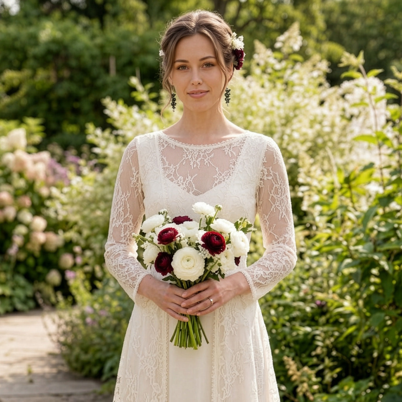 Bride standing in a blooming summer garden, wearing a long ivory lace wedding dress and holding a bouquet of white and deep burgundy flowers. Her hair is styled in a soft updo adorned with white blossoms and burgundy roses, and she wears delicate dark green floral earrings by byMinta. Sunlight filters through the garden, creating a romantic and natural atmosphere.