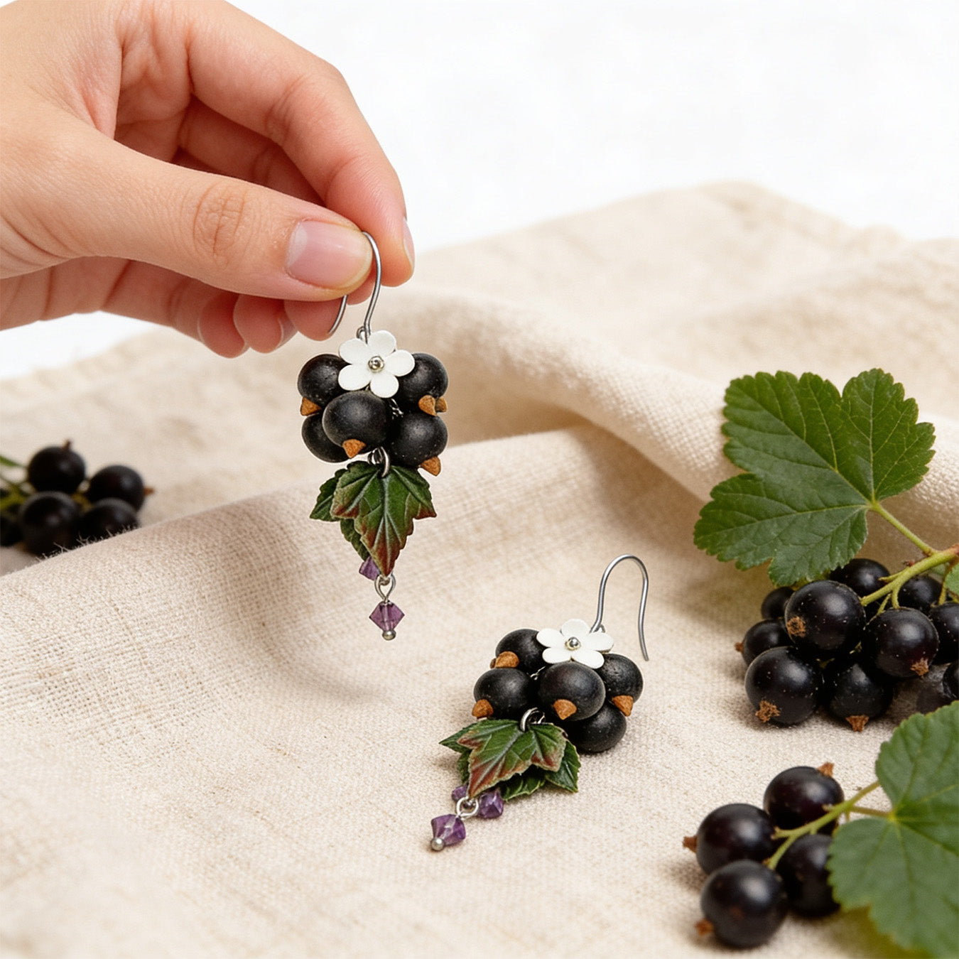 Shadow Currant Earrings photographed with fresh blackcurrants and green leaves in soft natural light, with one earring gently held by hand for scale. Handmade in Finland by byMinta.
