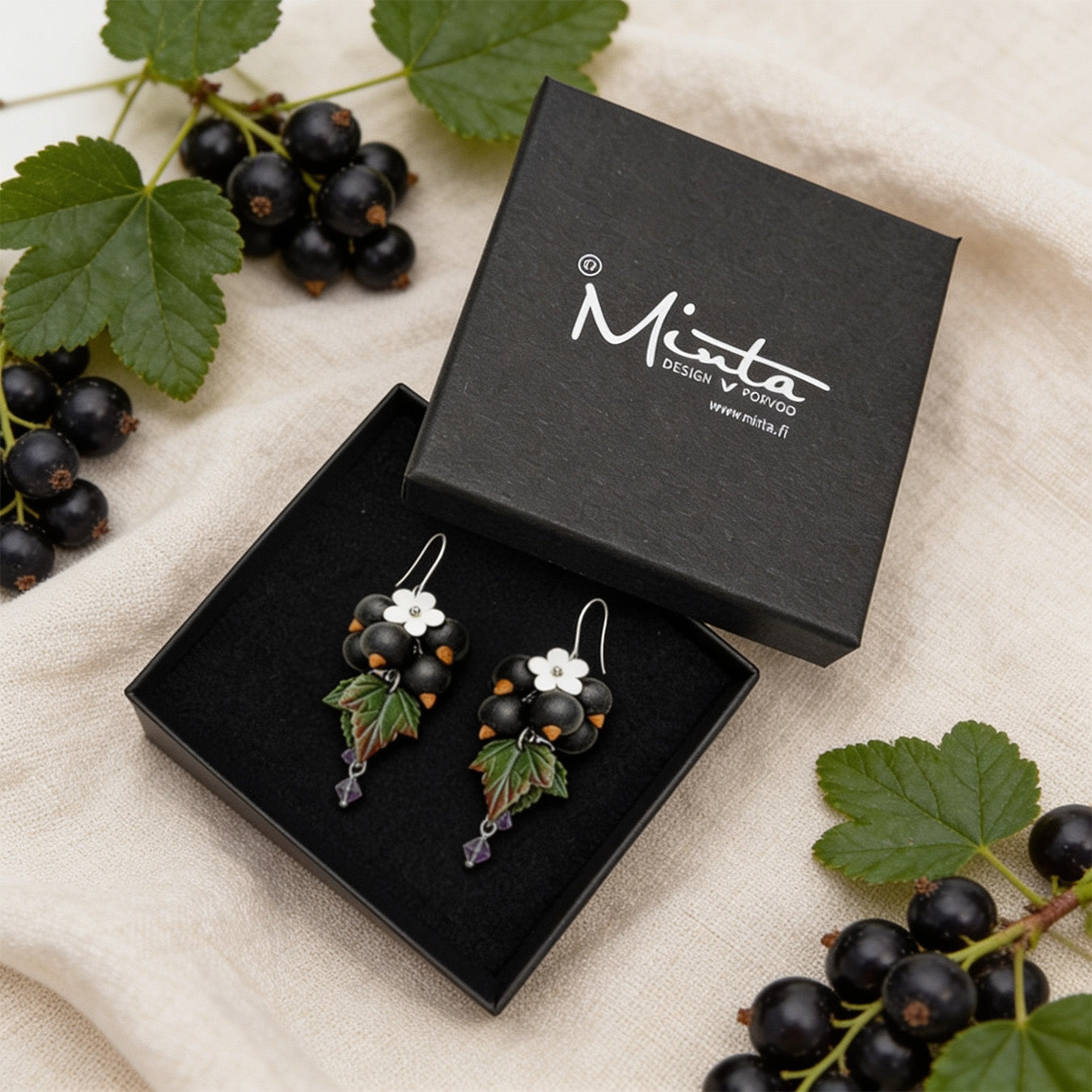 Shadow Currant Earrings presented in a gift box, surrounded by natural linen fabric, fresh blackcurrants, and green leaves. Handmade in Finland by byMinta.