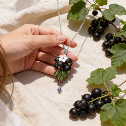 Shadow Currant Necklace held gently in hand for scale, featuring hand-shaped black currants and leaves made from polymer clay. Natural linen fabric, fresh black currants, and green currant leaves in soft natural light. Handmade in Finland by byMinta.