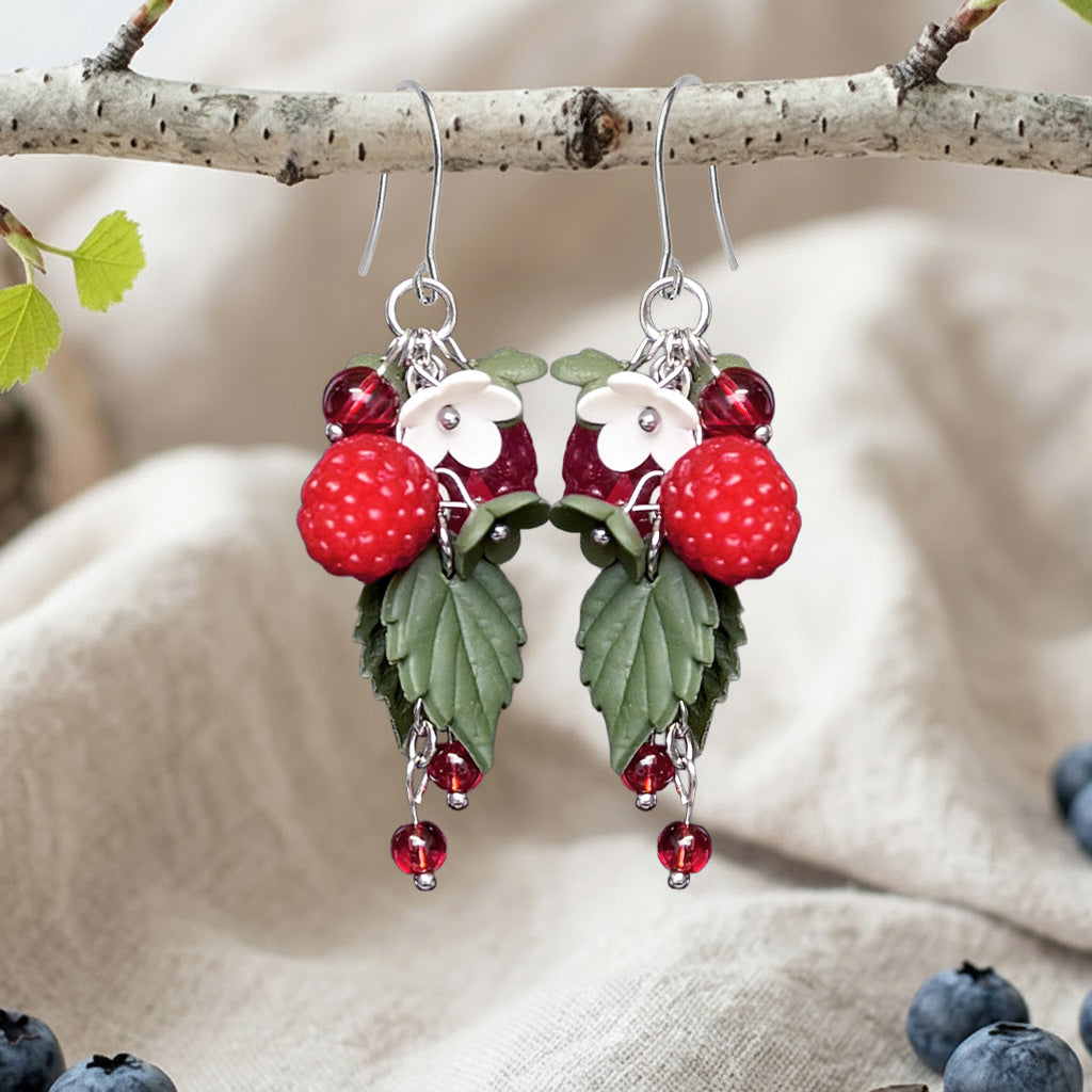 Handcrafted wild raspberry earrings hanging from a birch branch with white flowers and green leaves